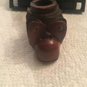 May include: A wooden pipe carved in the shape of a monkey's head. The pipe is brown and has a red nose and eyes.