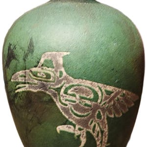 Puede incluir: Jarrón de cerámica verde con un diseño de ave estilizado y detallado en tonos grises. El jarrón tiene un borde oscuro y una superficie texturizada. El diseño del ave es una característica destacada, agregando un elemento artístico único.