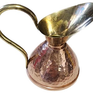 May include: A copper and brass pitcher with a hammered texture. The pitcher has a rounded base, a flared spout, and a curved handle. The handle and spout are brass, while the body is copper.