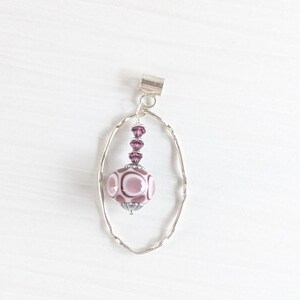 May include: A silver pendant featuring a large, round, pink and white bead. The bead is patterned and hangs within a silver oval frame. Above the bead are smaller purple beads and a silver clasp. The pendant is a piece of jewellery.