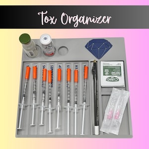 May include: A gray plastic Tox Organizer tray featuring the text Tox Organizer at the top. The tray holds seven syringes with orange caps, two small glass vials, a dark blue diamond-shaped inlay, a black marking pencil, and alcohol prep pads.