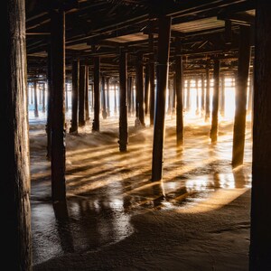 May include: A photograph taken from beneath a wooden pier, with sunlight streaming through the structure's supports. The scene is bathed in warm, golden light, creating long shadows on the wet sand below.