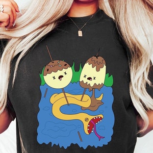 May include: Black t-shirt featuring a cartoon graphic of two chocolate-covered desserts with googly eyes and a snake with an open mouth. The text "Shroom Queens" is printed on the shirt.