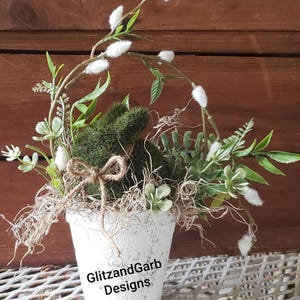May include: A white distressed pot with a moss-covered bunny and faux greenery. The arrangement includes white fluffy accents, green leaves, and a twine bow. The pot has the text "GlitzandGarb Designs" on the front.