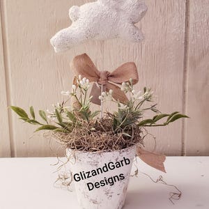 May include: A decorative Easter-themed arrangement in a distressed white pot. It features a white bunny figure, a brown bow, and small white flowers. The pot has the text "GlizandGarb Designs".