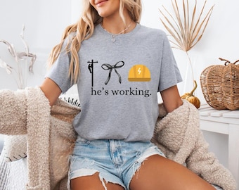 May include: A gray t-shirt with a black and white illustration of a person working on a power line, a black bow, and a yellow hard hat. The text "he's working." is printed below the illustration.