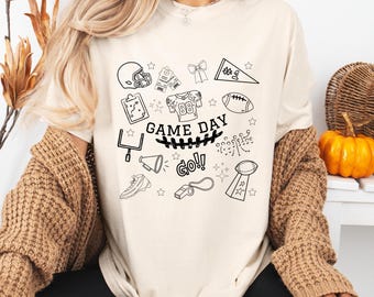 May include: Cream-colored t-shirt with black graphic illustrations of football-related items, including a helmet, tickets, and a football. The words "GAME DAY GO!" are prominently displayed. The shirt is part of a casual wear collection.
