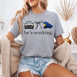 以下が含まれることがあります： 警察の車、青い警察の帽子、黒い蝶ネクタイがプリントされたグレーのTシャツ。Tシャツには「he's working.」という文字がプリントされています。