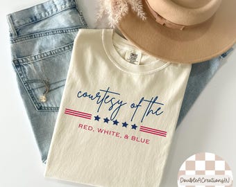 May include: Cream-colored t-shirt with the text "courtesy of the RED, WHITE, & BLUE" in navy and red. The shirt is laid on top of light-wash denim jeans, a beige hat, and a necklace.