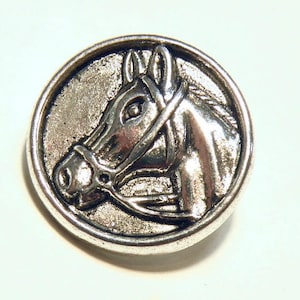 May include: Silver-toned metal button featuring a detailed horse head design. The horse is wearing a bridle and has a realistic look.