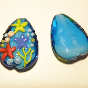 May include: Two glass beads, one with a blue and white swirl design and the other with a blue background and colorful sea life designs including starfish, coral, and seashells.