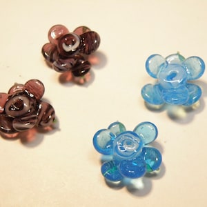 Puede incluir: Cuatro cuentas de vidrio con forma de flor, dos son azules y dos son moradas con remolinos blancos. Las cuentas tienen todas la forma de una flor de cinco p&eacute;talos.