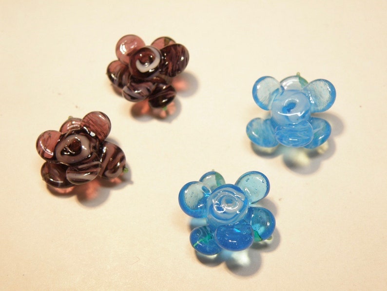 Puede incluir: Cuatro cuentas de vidrio con forma de flor, dos son azules y dos son moradas con remolinos blancos. Las cuentas tienen todas la forma de una flor de cinco p&eacute;talos.