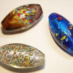 K&ouml;nnte beinhalten: Drei Glasperlen mit einem gesprenkelten Muster. Die Perlen sind oval geformt und haben eine Vielzahl von Farben, darunter Blau, Braun und Klar mit Silberflocken.