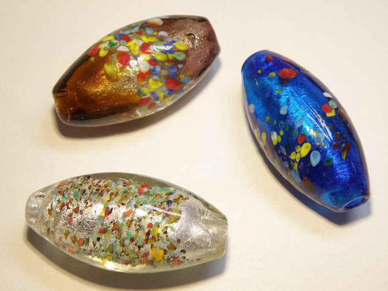 K&ouml;nnte beinhalten: Drei Glasperlen mit einem gesprenkelten Muster. Die Perlen sind oval geformt und haben eine Vielzahl von Farben, darunter Blau, Braun und Klar mit Silberflocken.