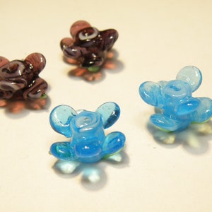 Puede incluir: Tres cuentas de flores de vidrio, una p&uacute;rpura con remolinos blancos, dos azul claro con remolinos blancos.