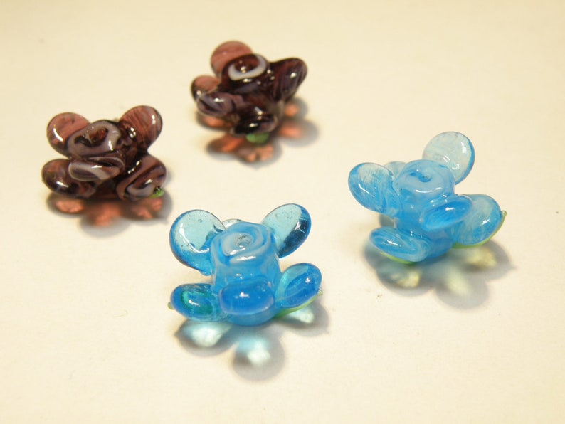 Puede incluir: Tres cuentas de flores de vidrio, una p&uacute;rpura con remolinos blancos, dos azul claro con remolinos blancos.