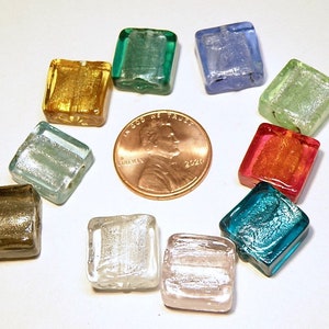 May include: A collection of colorful, square glass beads in various colors, including gold, green, blue, and red. A U.S. penny is in the center for size comparison. The beads have a textured, iridescent appearance.