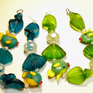 May include: Two strands of glass beads, one in teal and one in green, feature sea-themed charms, including seashells, fish, and swirls.