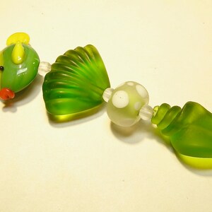 May include: A green glass bead necklace with a yellow and green fish charm, a green seashell charm, a white and green polka dot bead, and a green seashell charm.