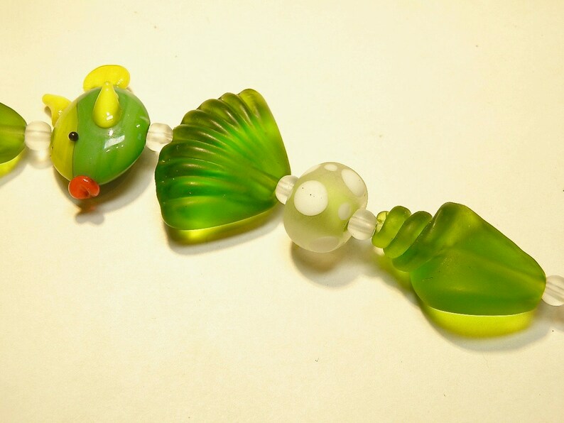 May include: A green glass bead necklace with a yellow and green fish charm, a green seashell charm, a white and green polka dot bead, and a green seashell charm.