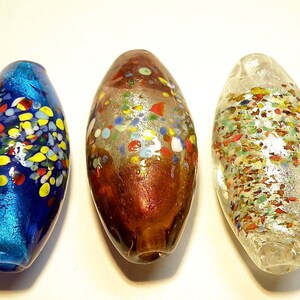 K&ouml;nnte beinhalten: Drei Glasperlen mit einem gesprenkelten Muster. Die Perlen sind oval geformt und haben eine blaue, braune und klare Glasbasis. Die Perlen sind mit bunten Glasflocken verziert.