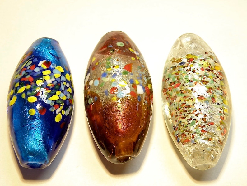 K&ouml;nnte beinhalten: Drei Glasperlen mit einem gesprenkelten Muster. Die Perlen sind oval geformt und haben eine blaue, braune und klare Glasbasis. Die Perlen sind mit bunten Glasflocken verziert.