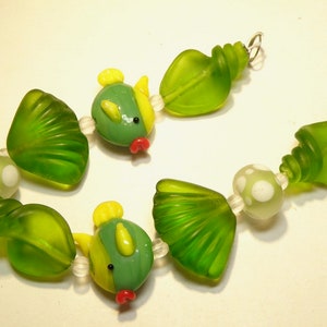 May include: A green and yellow glass bead necklace with a seashell and fish design. The necklace is made of glass beads and features a variety of shapes, including seashells, fish, and swirls.