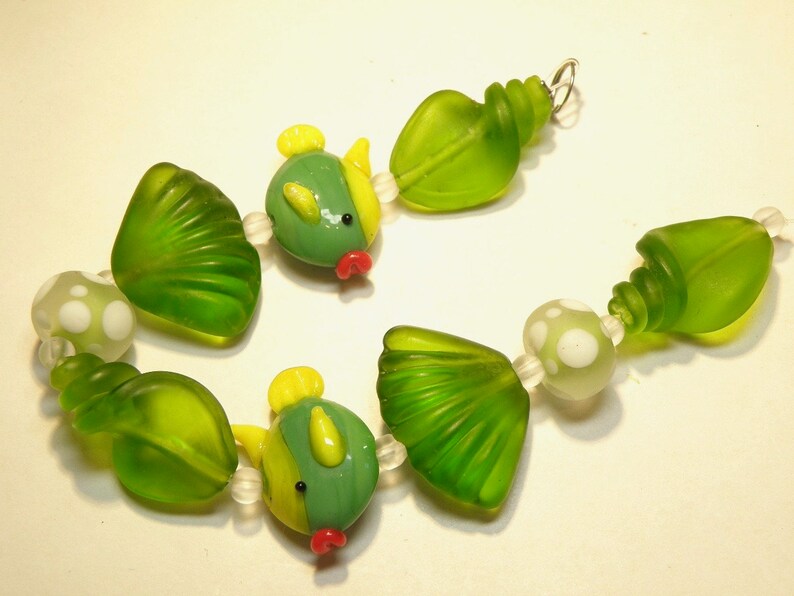 May include: A green and yellow glass bead necklace with a seashell and fish design. The necklace is made of glass beads and features a variety of shapes, including seashells, fish, and swirls.