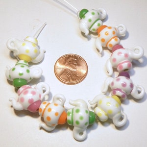 May include: A string of white teapot-shaped glass beads with colorful polka dots. The beads are strung on a white cord and are arranged in a circle.