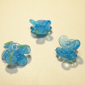 Puede incluir: Tres cuentas de vidrio azul con detalles verdes. Las cuentas tienen forma de flores con m&uacute;ltiples p&eacute;talos.