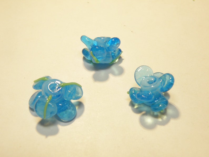 Puede incluir: Tres cuentas de vidrio azul con detalles verdes. Las cuentas tienen forma de flores con m&uacute;ltiples p&eacute;talos.