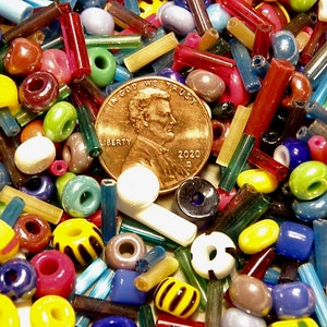 May include: A close-up image of a variety of colorful glass beads and glass tubes. The beads are in various shapes and sizes, including round, oval, and cylindrical. The tubes are also in various colors and lengths. A US penny coin is in the center of the image, with the text "In God We Trust" and "Liberty" visible. The coin is dated 2020.