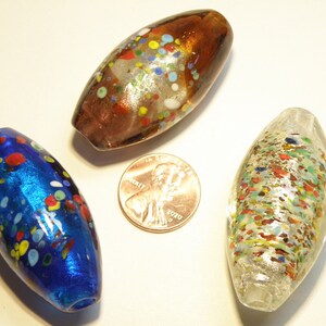 K&ouml;nnte beinhalten: Drei Glasperlen mit einem gesprenkelten Muster. Die Perlen sind oval geformt und haben einen metallischen Glanz. Die Perlen sind blau, braun und silber.