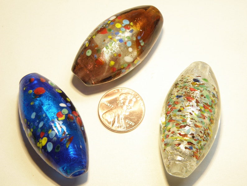 K&ouml;nnte beinhalten: Drei Glasperlen mit einem gesprenkelten Muster. Die Perlen sind oval geformt und haben einen metallischen Glanz. Die Perlen sind blau, braun und silber.
