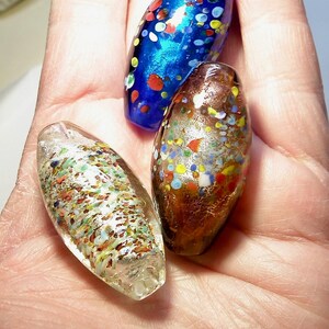K&ouml;nnte beinhalten: Drei Glasperlen mit einem gesprenkelten Muster. Die Perlen sind oval geformt und haben eine Vielzahl von Farben, darunter Blau, Gold und Wei&szlig;.