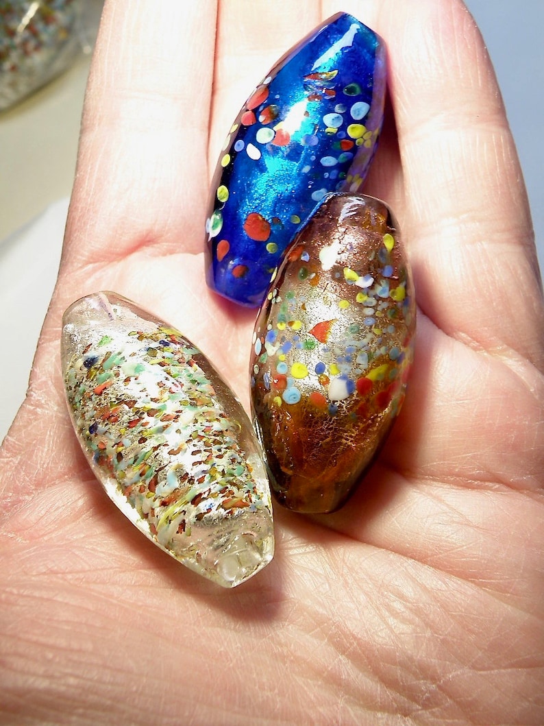 K&ouml;nnte beinhalten: Drei Glasperlen mit einem gesprenkelten Muster. Die Perlen sind oval geformt und haben eine Vielzahl von Farben, darunter Blau, Gold und Wei&szlig;.