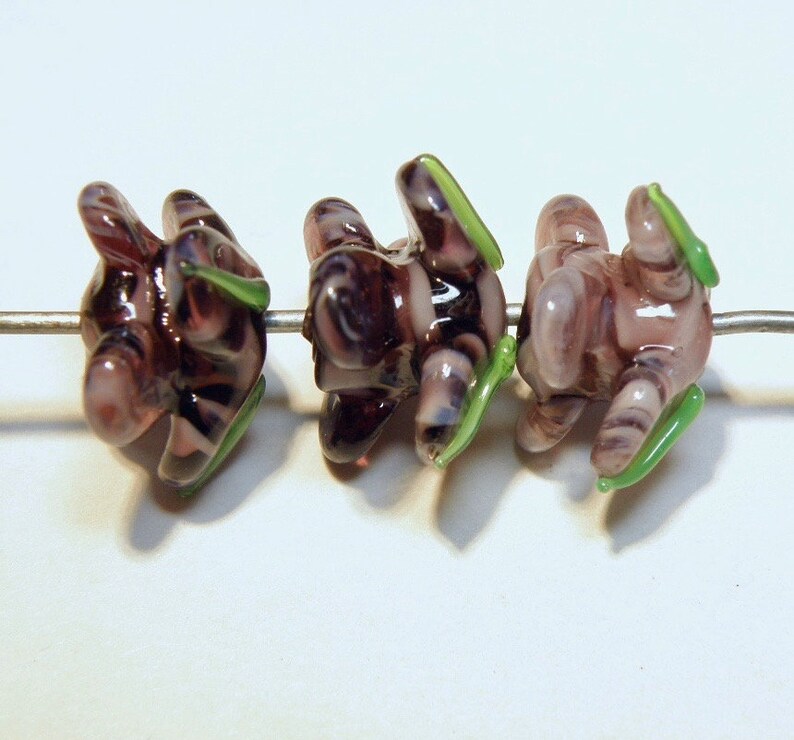 Puede incluir: Tres cuentas de vidrio con un patr&oacute;n de espiral morado y blanco y detalles verdes. Las cuentas est&aacute;n enhebradas en un alambre.