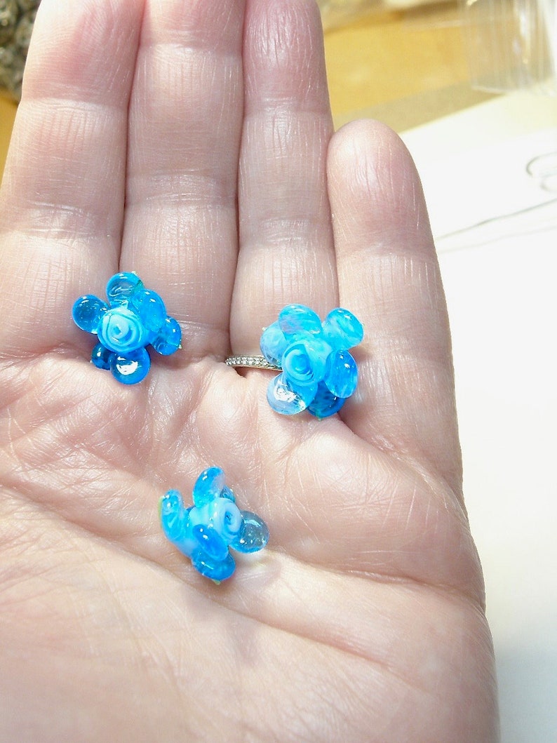 Puede incluir: Tres peque&ntilde;os abalorios de flores de vidrio azul y blanco. Los abalorios tienen la forma de una flor con cinco p&eacute;talos y un centro.