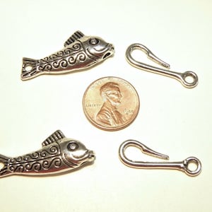 May include: Two silver fish charms with a swirl design and a silver hook clasp. The charms are about the size of a US penny.