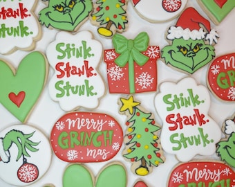 Puede incluir: Una colección de galletas decoradas con caras verdes de Grinch, cajas de regalo rojas con lazos verdes, corazones verdes con centros rojos, árboles de Navidad y placas blancas con el texto rojo y verde Stink. Stank. Stunk. o Merry Grinchmas.