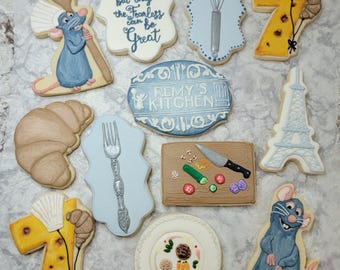 May include: A set of 10 decorated sugar cookies featuring characters and objects from the animated film Ratatouille. The cookies are decorated with intricate details and vibrant colors. The cookies include a rat chef, a whisk, a croissant, a fork, a cutting board with vegetables, a plate with food, a Eiffel Tower, a cheese wheel, and two cookies with the quote "Anyone can cook, but only the fearless can be great."