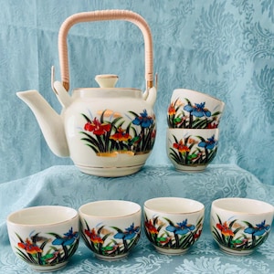 May include: A white ceramic tea set featuring a teapot with a woven handle and six matching teacups. The set is decorated with colorful floral designs in red, blue, and yellow, with gold trim. The teapot has a height of approximately 6 inches.