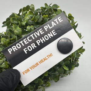Op de afbeelding: Een zwart-witte kaart met de tekst "Protective Plate for Phone" en "For Your Health!" Een zwarte ronde magneetplaat is aan de kaart bevestigd.