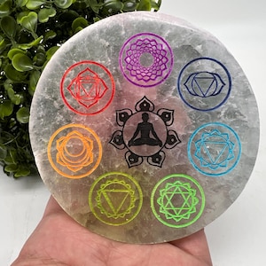Puede incluir: Un disco de cristal de selenita blanco con siete símbolos de chakras grabados en diferentes colores. Los símbolos están dispuestos en un círculo alrededor de una figura meditando.