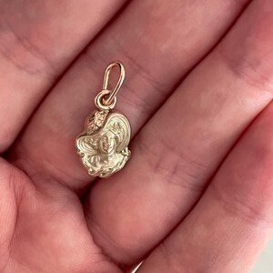 May include: A gold-colored pendant featuring a detailed relief of a woman's face wearing a hat. The pendant has a small loop at the top for attaching to a chain. The design is intricate and shows fine details.