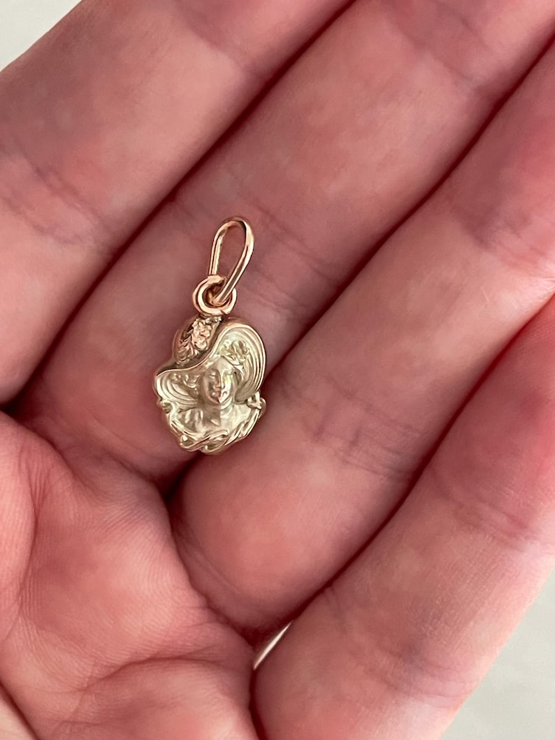 May include: A gold-colored pendant featuring a detailed relief of a woman's face wearing a hat. The pendant has a small loop at the top for attaching to a chain. The design is intricate and shows fine details.