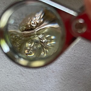 May include: Close-up of a gold-toned pendant with a detailed relief of a classical female face and ornate wings. The pendant is viewed through a magnifying glass, highlighting the intricate design and craftsmanship. The background is a soft, neutral color.