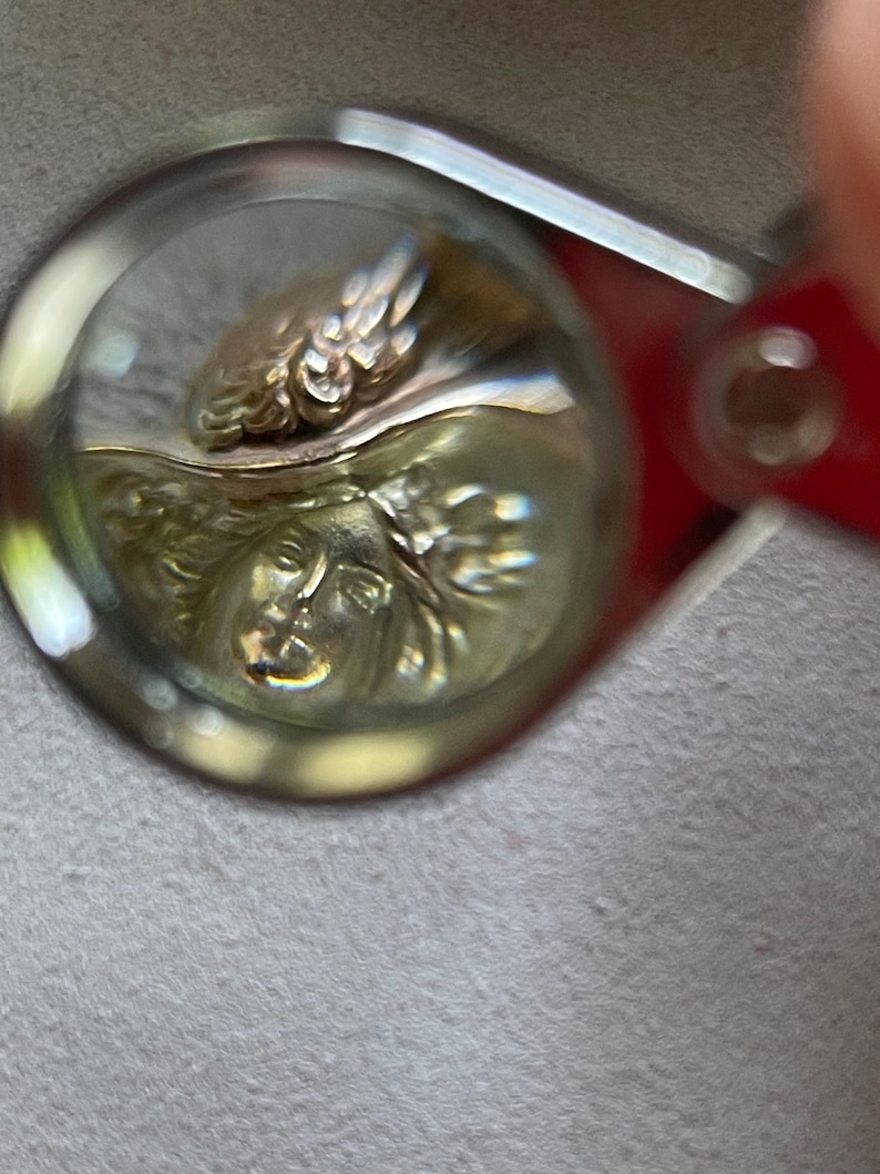 May include: Close-up of a gold-toned pendant with a detailed relief of a classical female face and ornate wings. The pendant is viewed through a magnifying glass, highlighting the intricate design and craftsmanship. The background is a soft, neutral color.