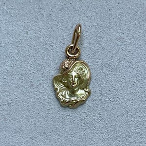May include: A gold-colored pendant featuring a woman's face wearing a wide-brimmed hat. The pendant has intricate details, including floral accents on the hat. The bail is a simple loop for attaching to a chain. The pendant is likely made of gold.